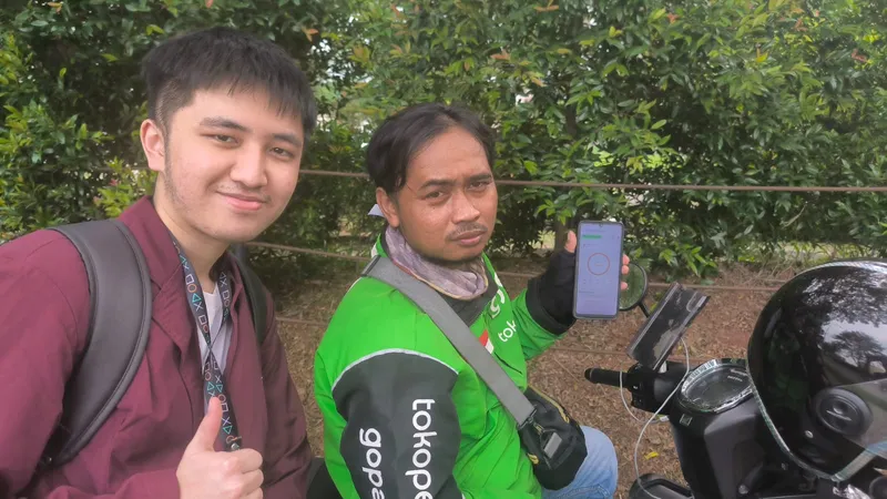 An online driver seated on his motorbike holding a phone with the app, next to a student giving a thumbs up